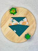 Overhead view of a reversible pet bandana with an orange and green citrus pattern on one side and dark teal with dots on the other. The bandana is on a round wooden surface with green succulents in white pots at the top left and bottom right.