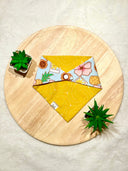 Overhead view of a reversible pet bandana. One side has a tropical print on light blue fabric, the other is yellow with white dotted swirls. The bandana is on a round wooden surface with green succulents in pots at the top left and bottom right.