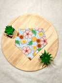 Overhead view of a light blue pet bandana with a tropical print of pink hibiscus flowers, pineapples, and postage stamps. The bandana is on a round, light wooden surface with green succulents in white pots at the top left and bottom right.