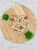 Overhead view of a pet bandana with a whimsical forest print featuring mushrooms, tree stumps, flowers, and woodland creatures on a beige background. The bandana is neatly laid out, showcasing its charming and detailed design.
