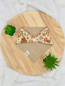 Overhead view of a reversible pet bandana featuring a whimsical forest print with mushrooms, tree stumps, flowers, and woodland creatures on one side and a burlap-like beige fabric on the other. The bandana is neatly laid out, showcasing both sides.